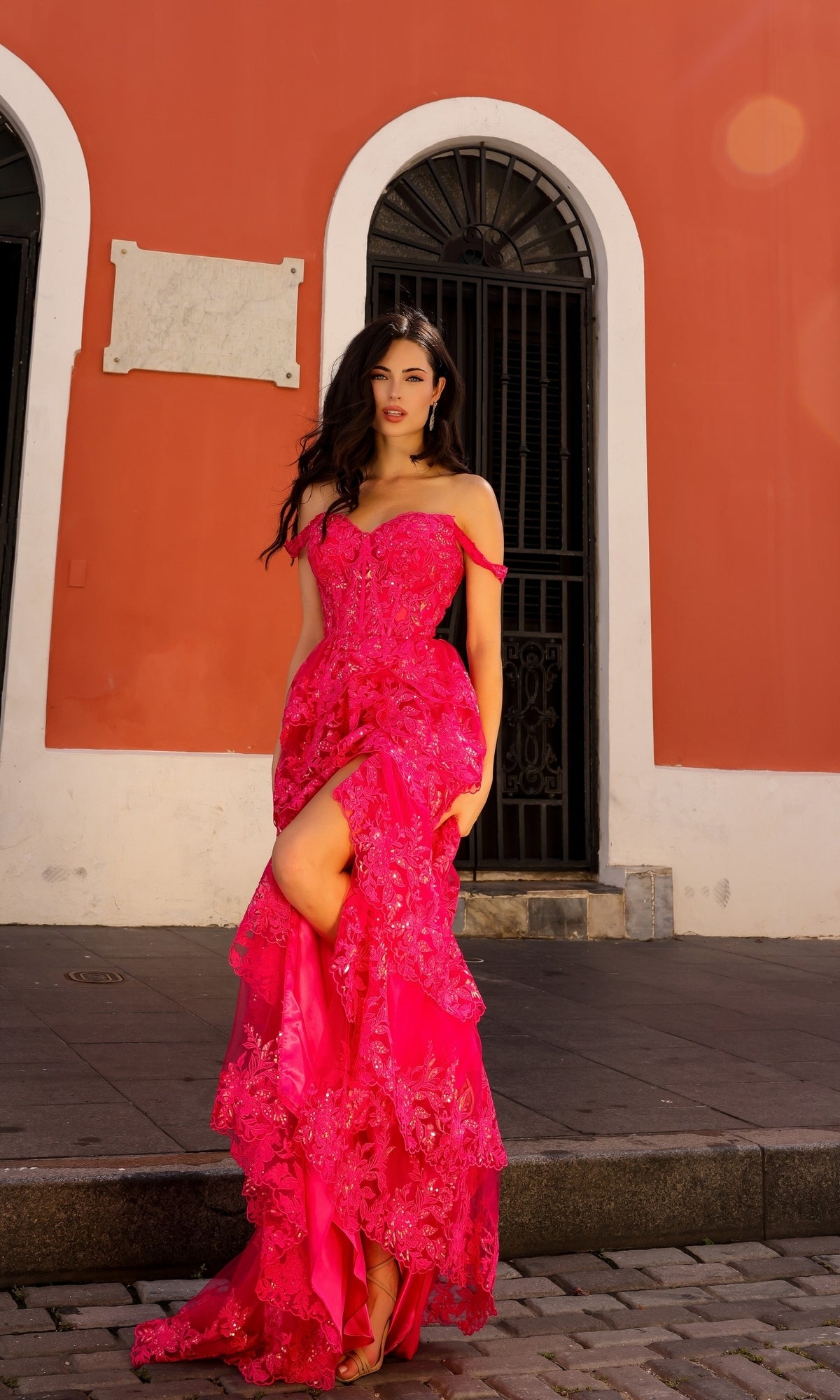 A woman in the Long Lace Ruffled A-Line Prom Dress T1335, featuring a bright pink off-shoulder design, stands on a cobblestone street before a red and white building with arched windows, ready for formal events.